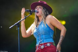 Female singer performing on stage, wearing a maroon hat and denim top, singing into a mic with a tattooed arm visible on the right.