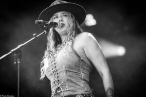 Female singer in a wide-brim hat performs into a microphone on stage, wearing a lace-up denim top with visible tattoos on her arm (black-and-white).