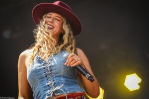 Smiling female singer wearing a burgundy hat and lace-up denim top, holding a microphone on stage.