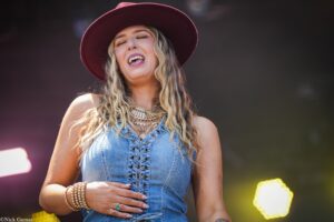Woman wearing a burgundy hat and denim top sings with eyes closed on stage outdoors, wearing layered jewelry.