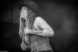 Female singer on stage, wearing a wide-brim hat and denim vest, smiling while holding a microphone with tattooed arm visible on a dark background.