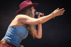 Female singer wearing a burgundy hat and denim vest, microphone in hand, reaching out to the audience with bracelets on her wrist.