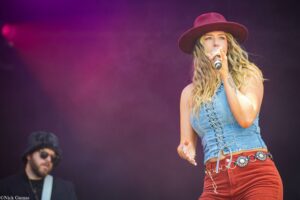 Female singer on stage, wearing a burgundy hat and denim top, singing into a microphone with purple stage lighting behind her.
