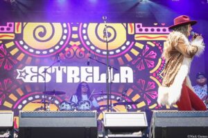 Female singer in a brown fur coat and red hat performs into a microphone on a colorful stage backdrop, drummer visible in the background with the word 'ESTRELLA' on the banner.
