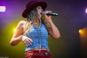 Female singer in a denim sleeveless top and maroon hat singing into a microphone on stage with purple and yellow lights behind her.