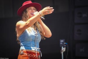 Female singer on stage, wearing a red wide-brim hat, denim vest, and orange belt, mid-performance into a microphone callout.