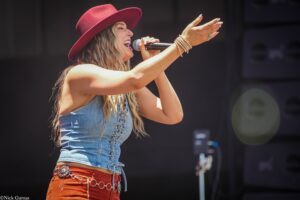 Female singer wearing a red hat and denim vest sings into a microphone on stage, arms raised toward the audience.