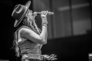 Female singer on stage, wearing a wide-brim hat and denim outfit, singing into a microphone with braced bracelet-clad arms visible.