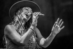 Female singer performing on stage into a microphone, wearing a wide hat and stacked bracelets, with hands raised in song (black and white).