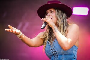 Female singer performing on stage, wearing a burgundy hat and denim vest, singing into a microphone while gesturing outward.