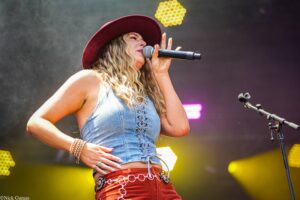 Female singer performing on stage with a denim top and red hat, holding a microphone as she sings; yellow stage lights in the background.