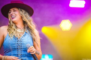 Female singer wearing a denim vest and brown hat, laughing while holding a microphone on a colorful stage backdrop.