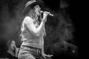 Female singer wearing a wide-brim hat and denim outfit, singing into a microphone on a smoky stage.