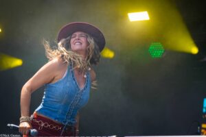 Female singer on stage wearing a denim top and wide hat, holding a microphone amid yellow stage lights.