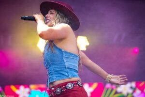 Female singer on stage, wearing a burgundy hat and denim top, singing into a handheld mic with colorful lights behind.