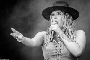 Female singer wearing a wide-brimmed hat performs into a microphone, reaching toward the audience on a smoky stage (black and white).