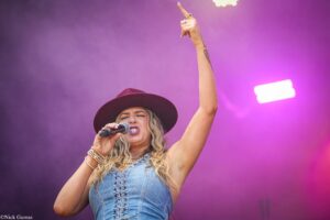 Female singer on stage loudly singing into a handheld mic, right arm raised high, wearing a burgundy hat and denim top against purple stage lighting.
