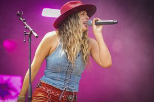 Female singer on stage wearing a red hat and denim top, singing into a handheld microphone with purple lighting in the background.