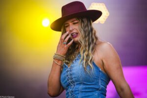 Woman in a burgundy wide-brim hat wearing a denim sleeveless top, touching her face against a colorful stage backdrop with yellow and purple lights.