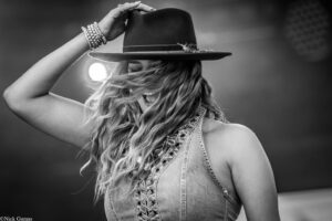 Person wearing a sleeveless denim vest, tipping a wide-brim hat while wind-tousled hair blows across the face.