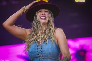 Woman on stage wearing a maroon hat and denim top, smiling with long wavy blonde hair, purple stage lights behind.