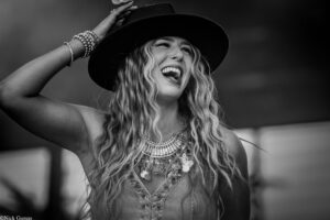 Joyful woman in a wide-brim hat laughs, touching the hat with her hand, wearing layered bracelets and a beaded necklace.