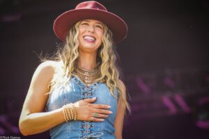 Smiling woman with long blonde hair wearing a maroon hat and denim top, hands resting on her chest outdoors.
