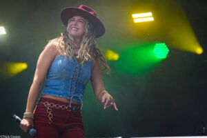 Female singer on stage wearing a burgundy wide-brimmed hat, denim top, red shorts, holding a microphone with colorful stage lights in the background.