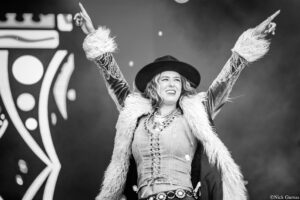 Black-and-white photo of a female performer on stage with arms raised, wearing a black hat and a fluffy fur coat.