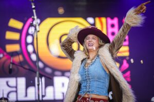 Joyful female performer on stage, raising arms in a burgundy hat and denim outfit with a fur vest.