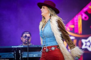 Female singer on stage wearing a burgundy wide-brim hat, denim vest with faux fur, red pants, holding a microphone amid purple stage lights.