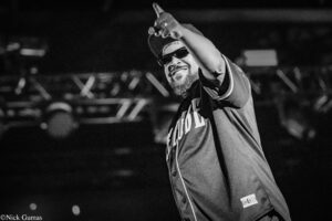 Concert performer on stage, wearing a cap and sunglasses, pointing upward to the crowd with a beaming smile in a black-and-white shot.
