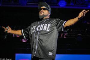 Performer on stage with arms wide, wearing a black baseball-style jersey, sunglasses, and a cap, holding a microphone in one hand against a dark background with blue stage lighting.