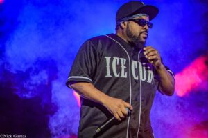 Male performer on stage, wearing sunglasses and a black baseball-style jersey, holding a microphone against blue-purple stage lighting.