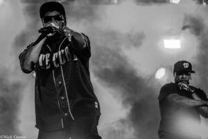 Male rapper wearing a cap and sunglasses performs into a microphone on a smoky stage.