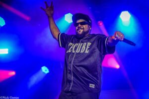 Male performer on a blue-lit stage, wearing sunglasses and a cap, holding a microphone with his right hand and raising his left hand in the air.