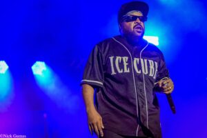 Ice Cube performing on stage in a dark jersey with white piping, wearing sunglasses and a head wrap, holding a microphone under blue stage lights.