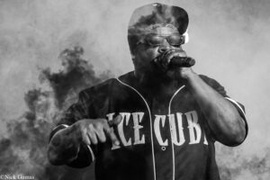 Black-and-white image of a male singer or rapper performing into a handheld microphone, wearing sunglasses, a backward cap, a bandana, and a jersey with 'NEW YORK' lettering, with smoke in the background.