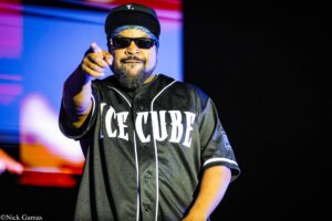 Hip-hop performer on stage wearing a black jersey with white piping and 'ICE CUBE' lettering, black cap, sunglasses, and bandana, pointing at the camera.