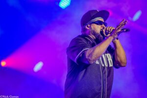 Male vocalist wearing a cap and sunglasses, singing into a handheld mic on a neon blue‑purple stage.