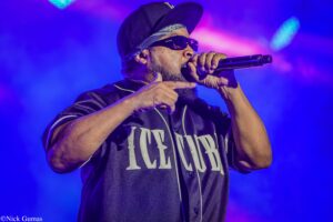 Male rapper performing on stage, mic in hand, wearing sunglasses, a dark cap, and bandana under blue stage lights.