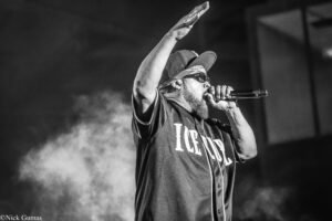 Black-and-white photo of a performer on stage, singing into a microphone with one arm raised above his head, wearing a cap and sunglasses.