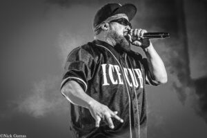 Black-and-white photo of a bearded male rapper on stage, wearing a cap and sunglasses, singing into a handheld microphone.