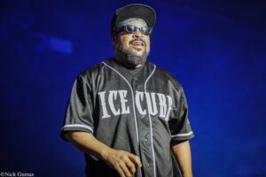 Hip-hop artist Ice Cube performing on stage in a black jersey with white piping and sunglasses against a blue background.”](text)