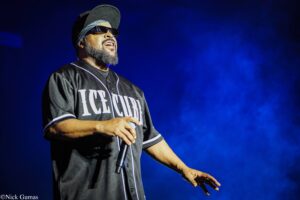 Male performer on stage wearing a black ICE CUBE jersey, cap, and sunglasses, holding a microphone with blue stage lighting behind him on the right side of the frame.