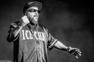 Black-and-white photo of a male rapper on stage, wearing a cap and sunglasses, singing into a microphone.