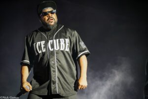 Ice Cube performing on stage in a black jersey with 'ICE CUBE' across the chest, holding a microphone.