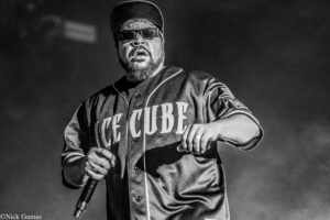 Male hip-hop performer on stage, wearing a cap and sunglasses, holding a microphone as he sings or raps.
