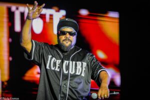 Male rapper on stage, wearing a black baseball jersey, cap, and sunglasses, waving his left hand and holding a microphone.