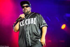Male performer on stage, holding a mic, wearing a black baseball jersey and sunglasses under red and blue lights.